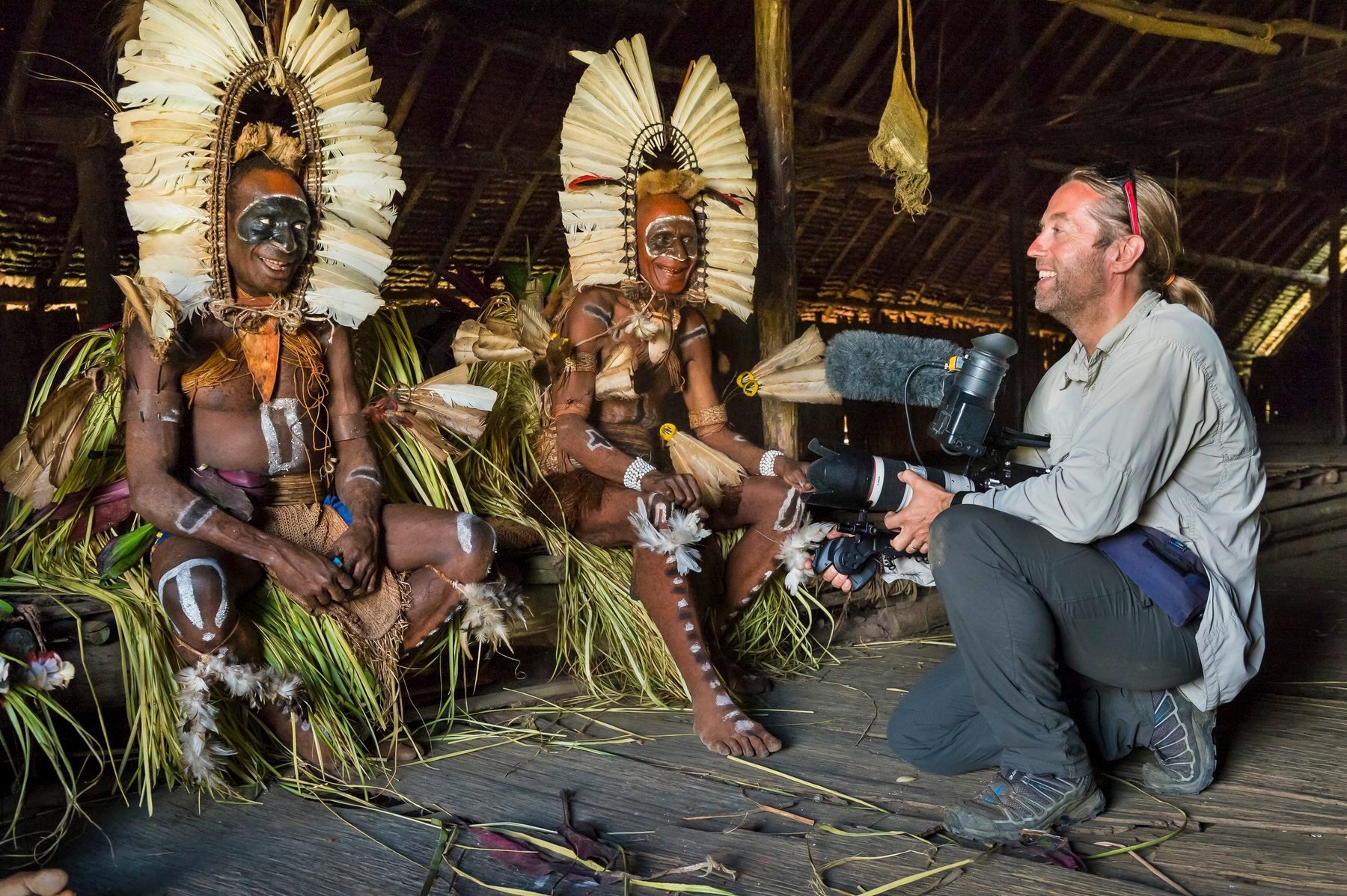 Cameraman filming two elders in ceremonial feather headdresses inside a men's house