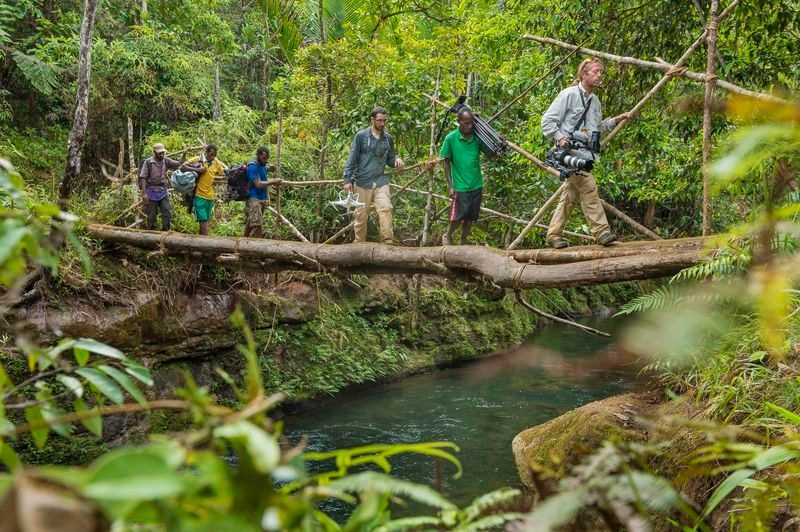 Film crew and porters crossing a log bridge over a jungle river