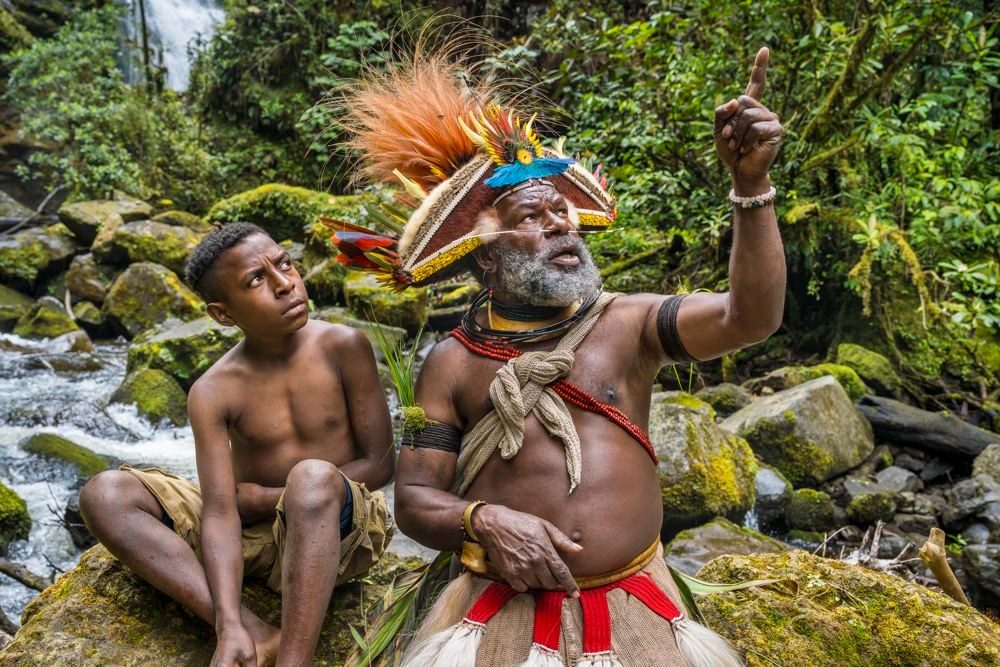 Huli elder in feather headdress pointing, beside a boy, near a river