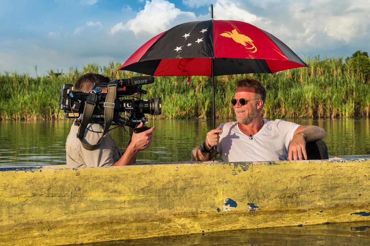 Cameraman filming a presenter under a Papua New Guinea flag umbrella in a canoe